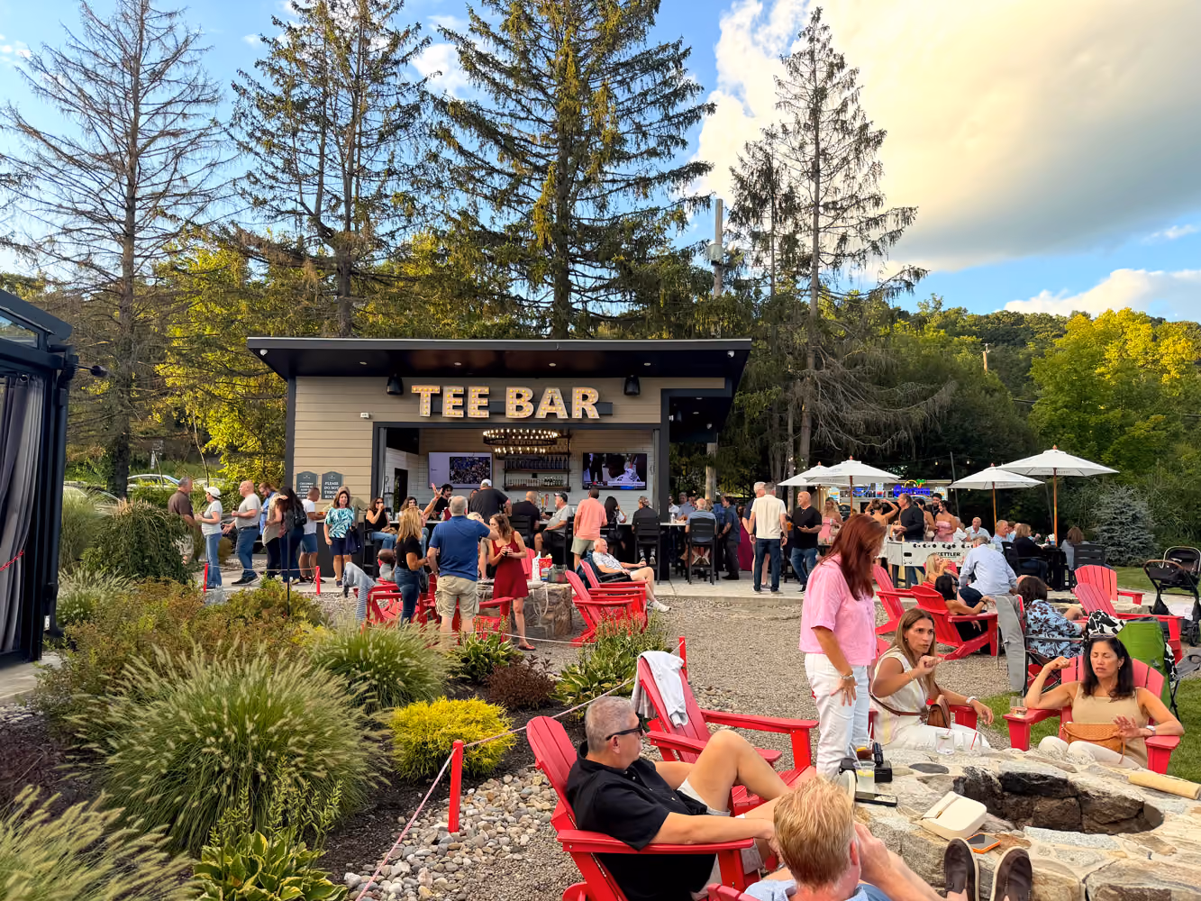 Outdoor bar named Tee Bar with people socializing and sitting on red chairs around fire pits.