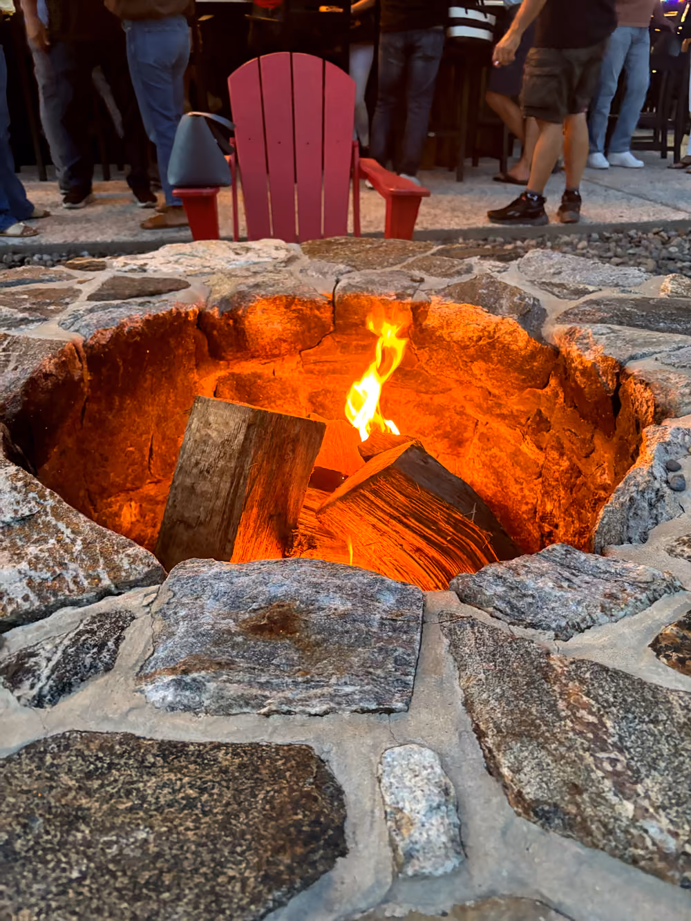 Stone fire pit with burning logs and a red Adirondack chair behind it outdoors.