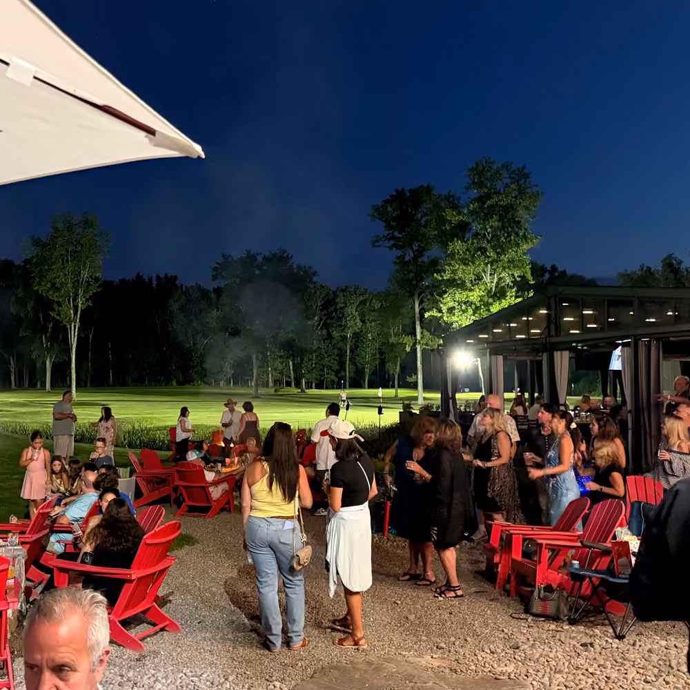 People socializing outdoors at night with red chairs, trees, and a lit pavilion nearby.