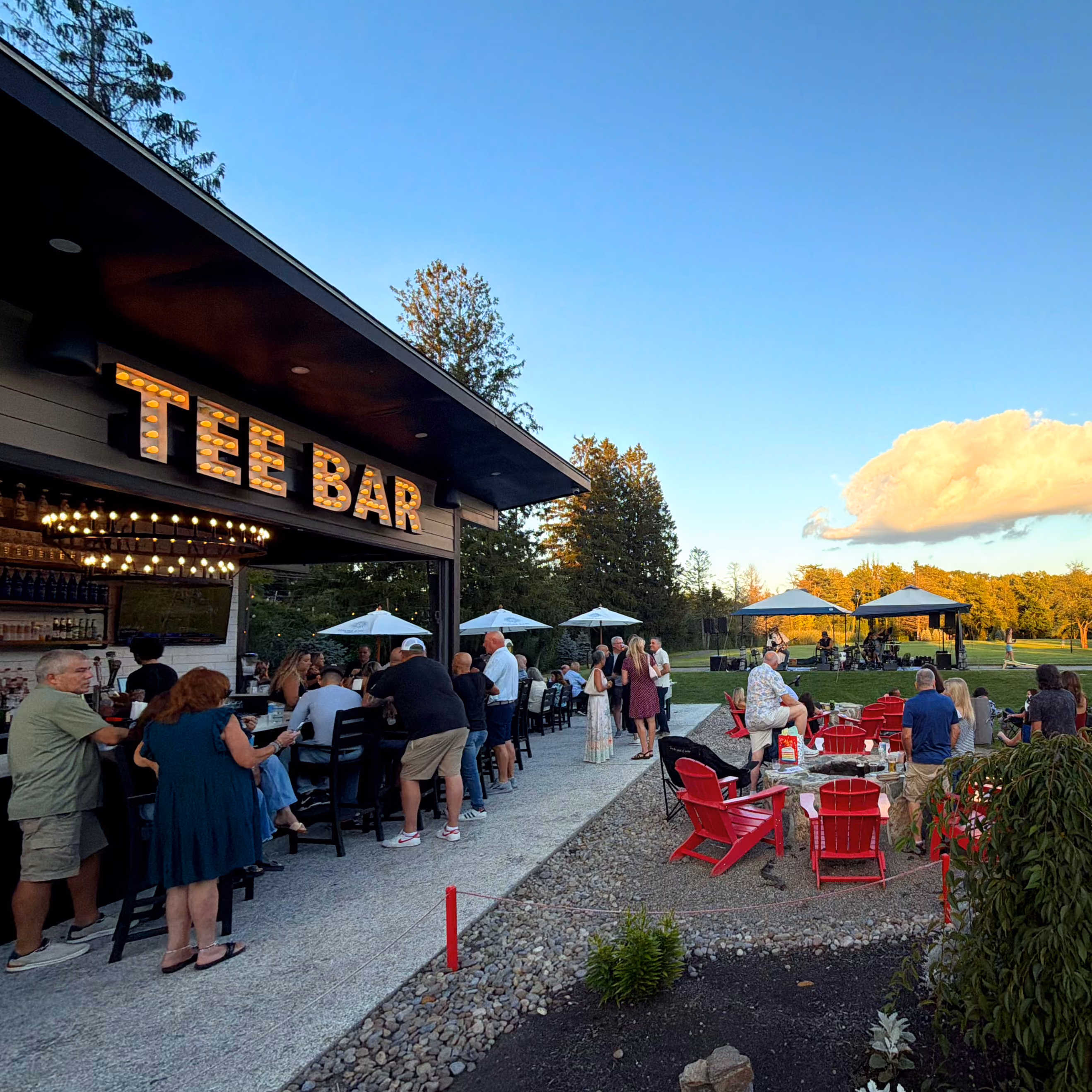 People gathered at an outdoor tee bar and seating area with umbrellas, red chairs, and a small live band.