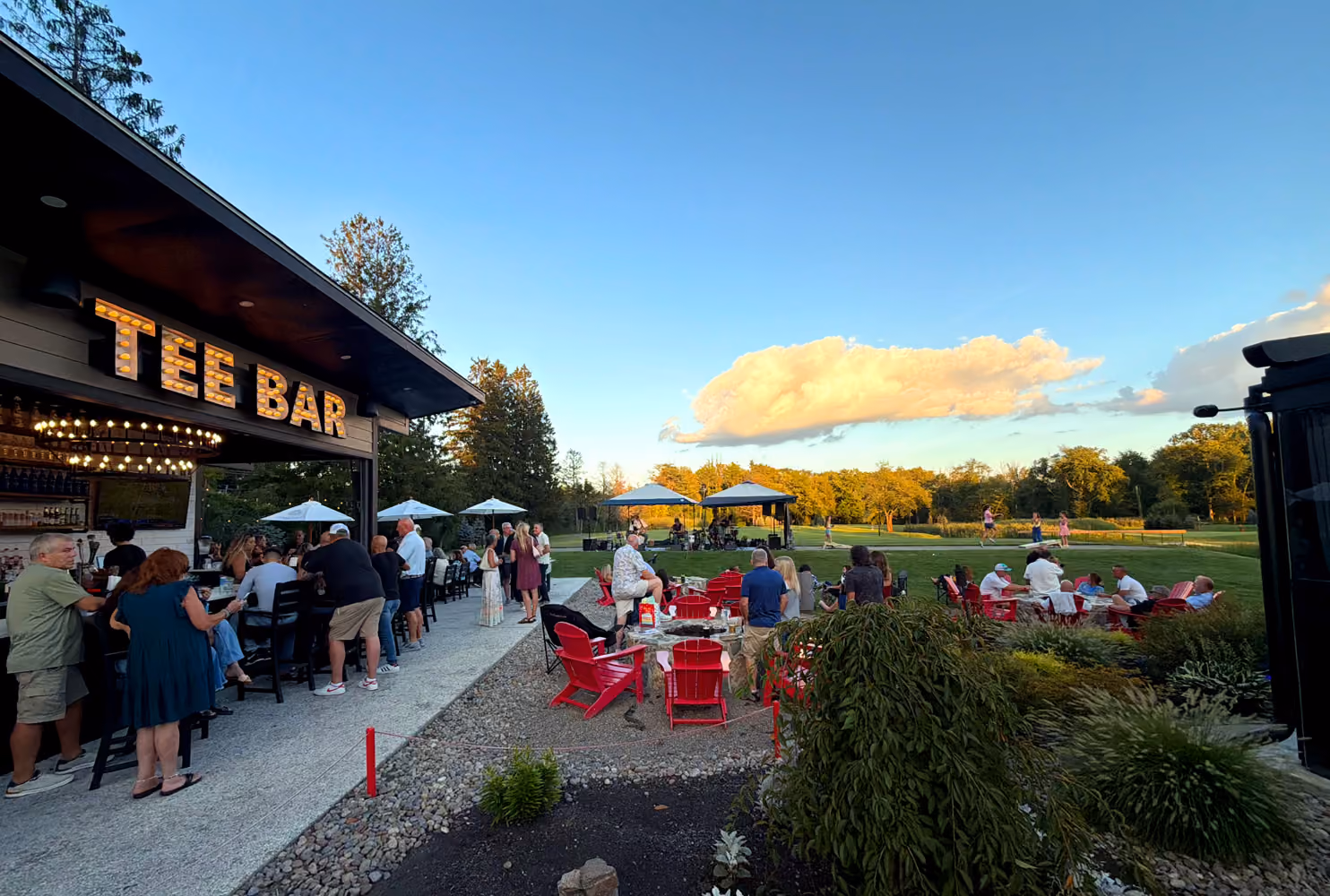 People socializing outside Tee Bar with red chairs and green lawn under blue sky with clouds.