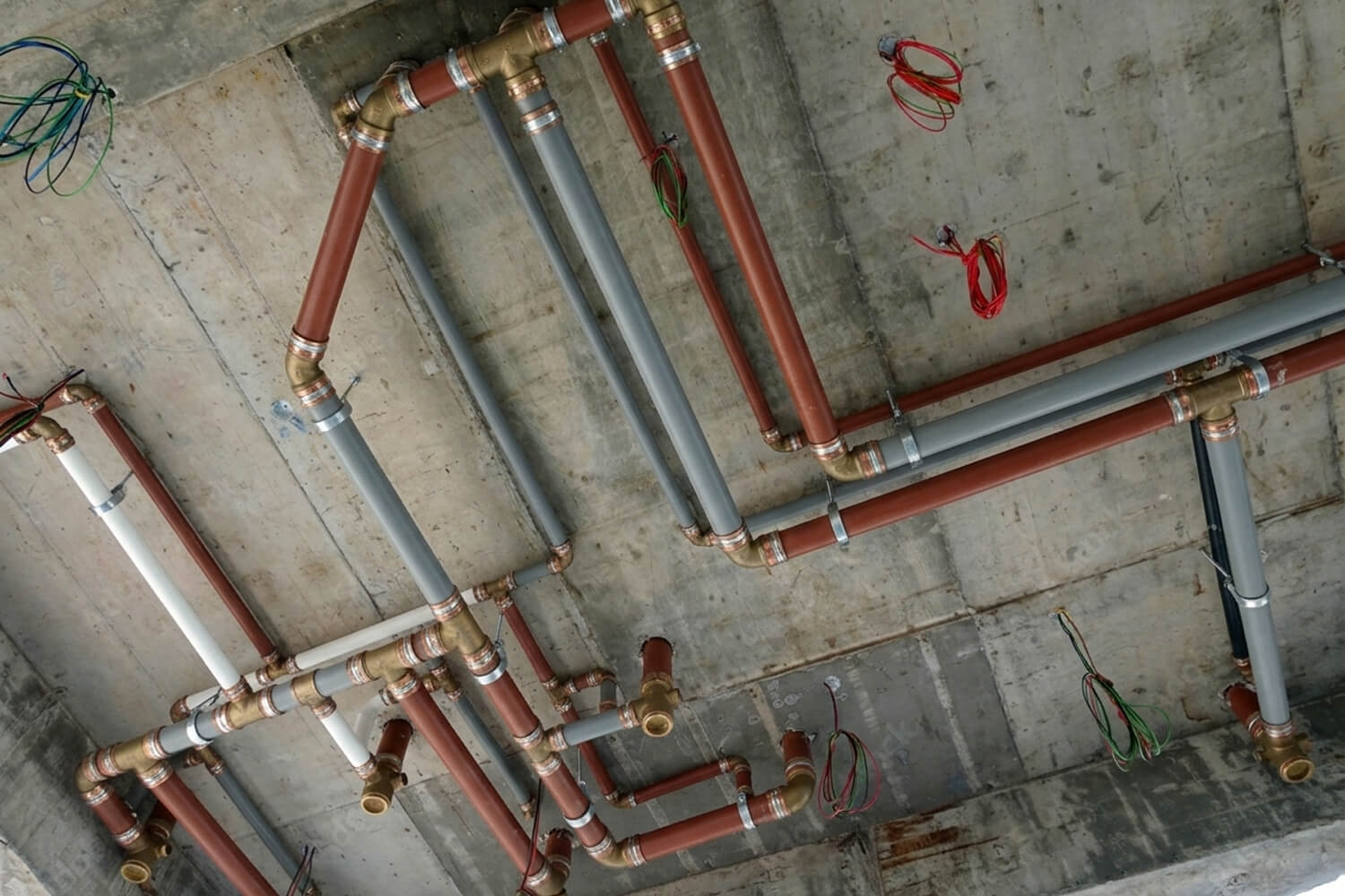 Complex ceiling-mounted plumbing system featuring insulated red and grey pipes with professional brass fittings and organized electrical wiring on concrete.