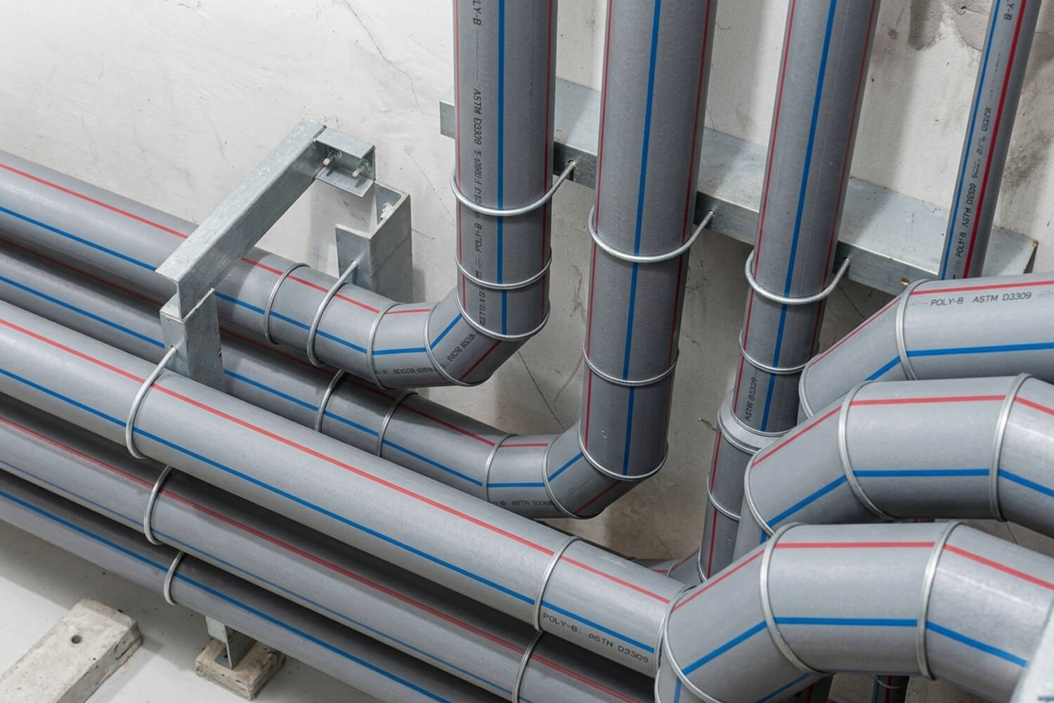 Close-up of industrial grey plumbing pipes with red and blue stripes organized with metal brackets against a concrete wall.