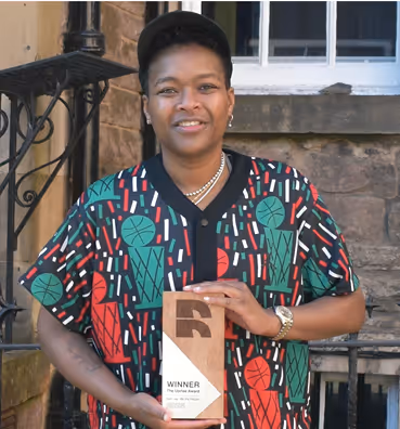 Person in a colorful geometric-patterned shirt and cap smiles, holding a winner's trophy. They stand in front of a stone building with a wrought iron railing.