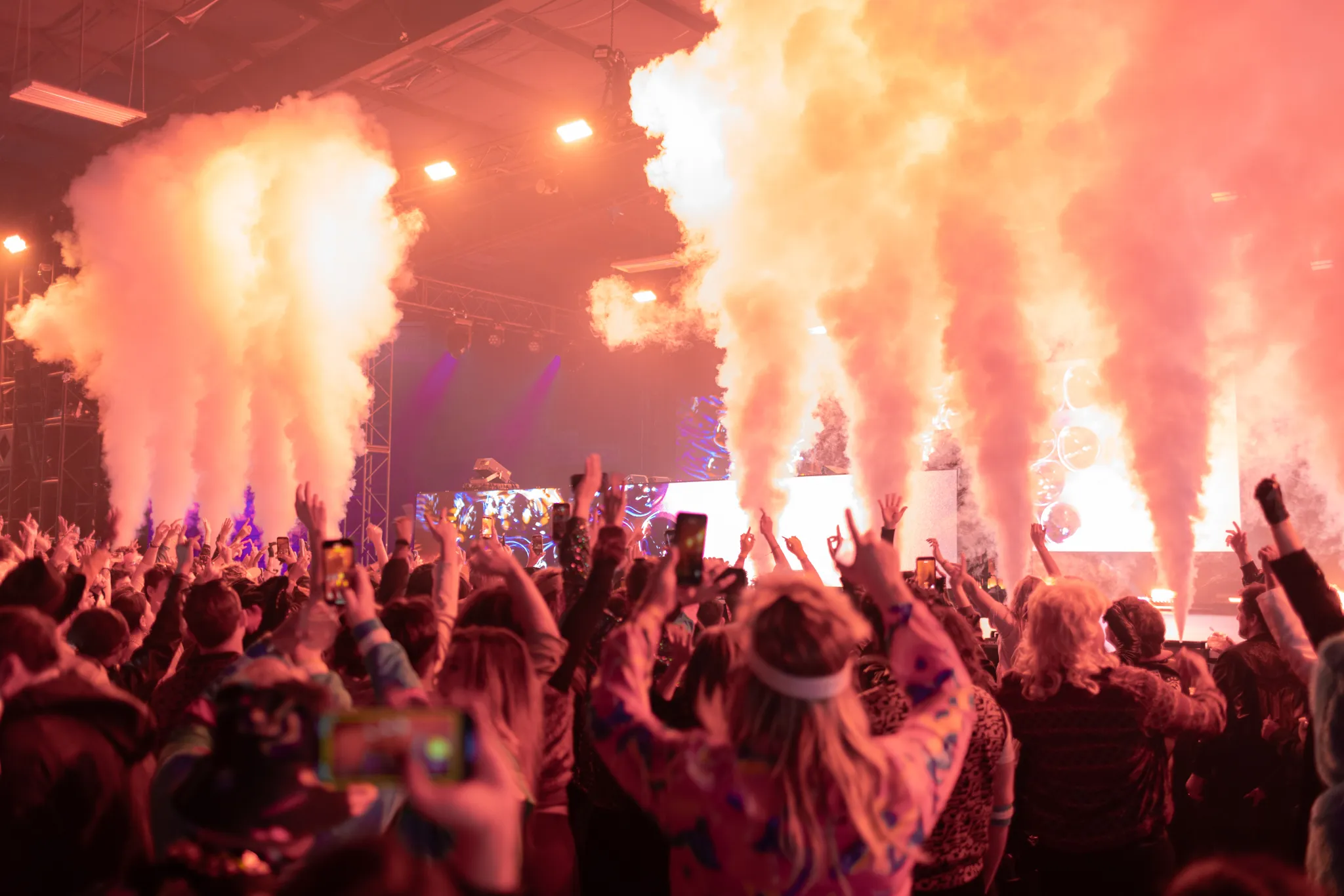 Crowd at a concert raising hands and phones while smoke effects rise from the stage.