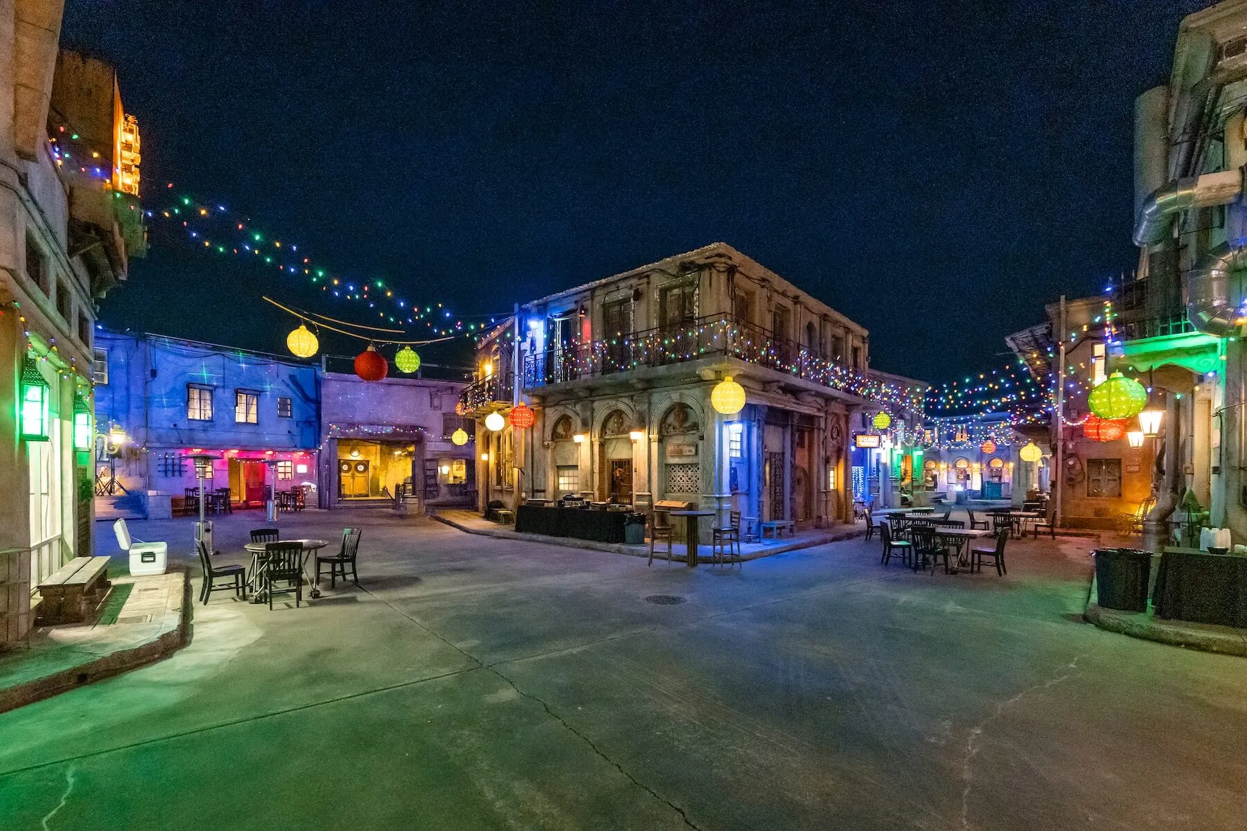 Nighttime street scene with old buildings decorated with colorful string lights and lanterns, with empty tables and chairs on the concrete ground.