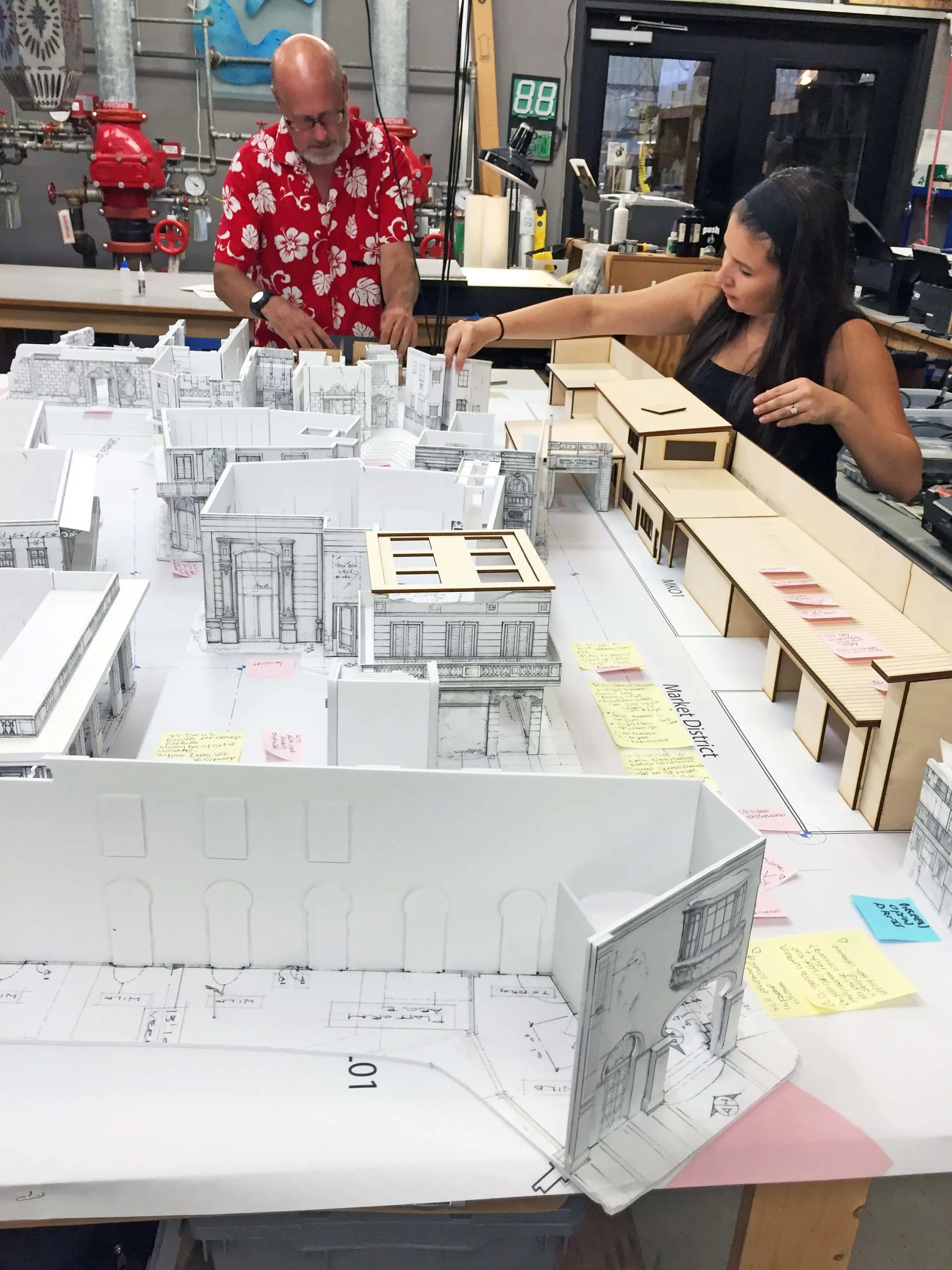 Two people working on an architectural model of a city layout with miniature buildings and sticky notes.