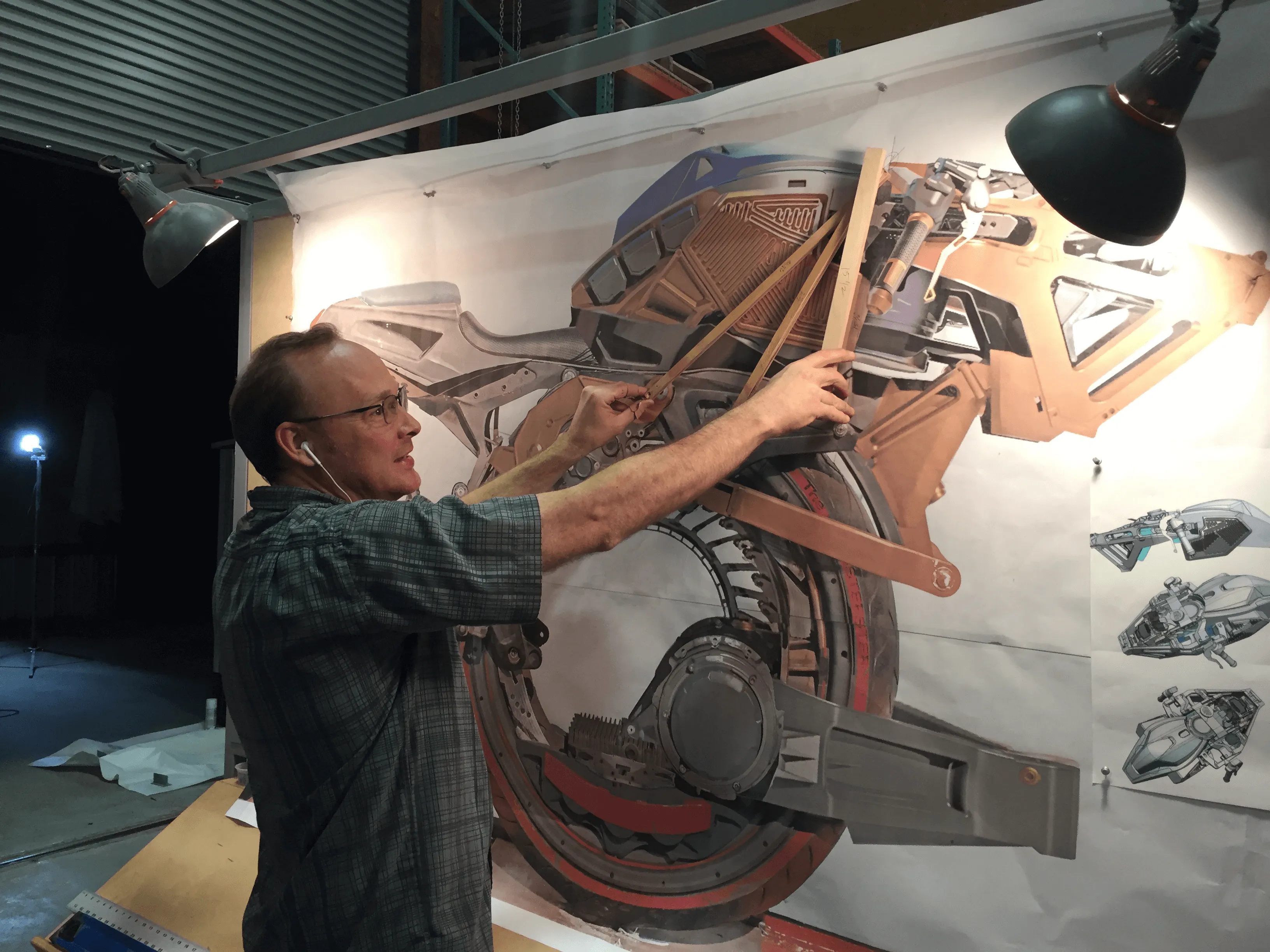 Man using wooden rulers to measure a large motorcycle design poster pinned on a wall under two lamps.