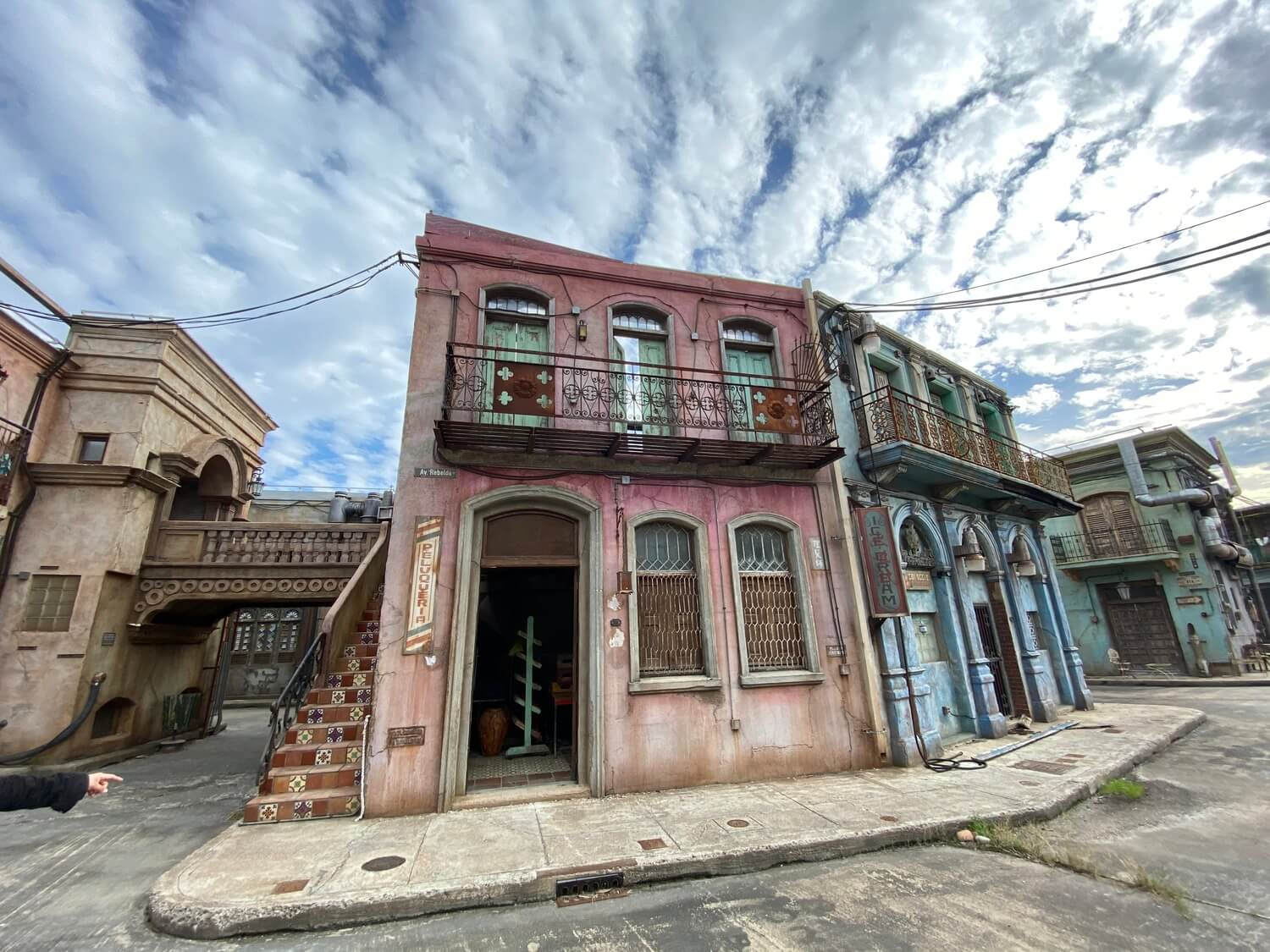 Street view of old buildings with weathered facades, one pink with a tiled staircase and a balcony, another blue with decorative columns and balconies, under a cloudy sky.