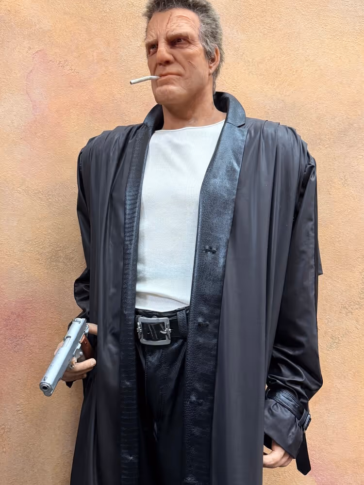 Realistic statue of a man with scars smoking a cigarette, wearing a black leather coat and holding a silver gun.