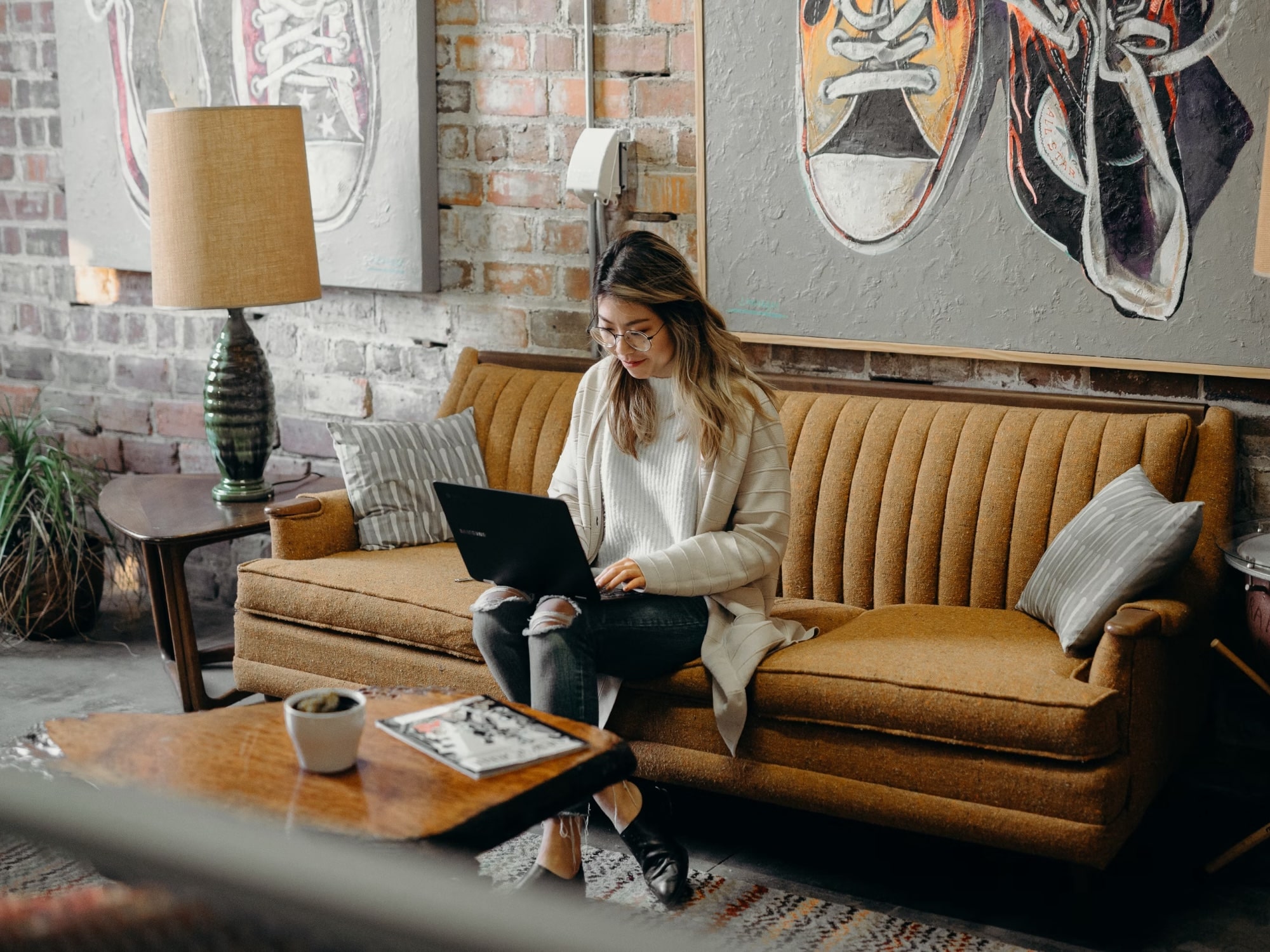 Woman with glasses sitting on a mustard yellow couch working on a laptop in a room with brick walls and colorful sneaker paintings.