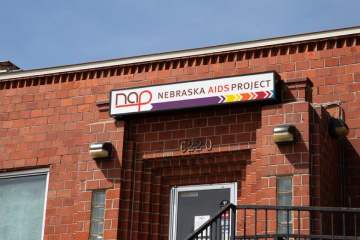 Brick building facade with a sign reading 'Nebraska AIDS Project' above a door under a clear sky.