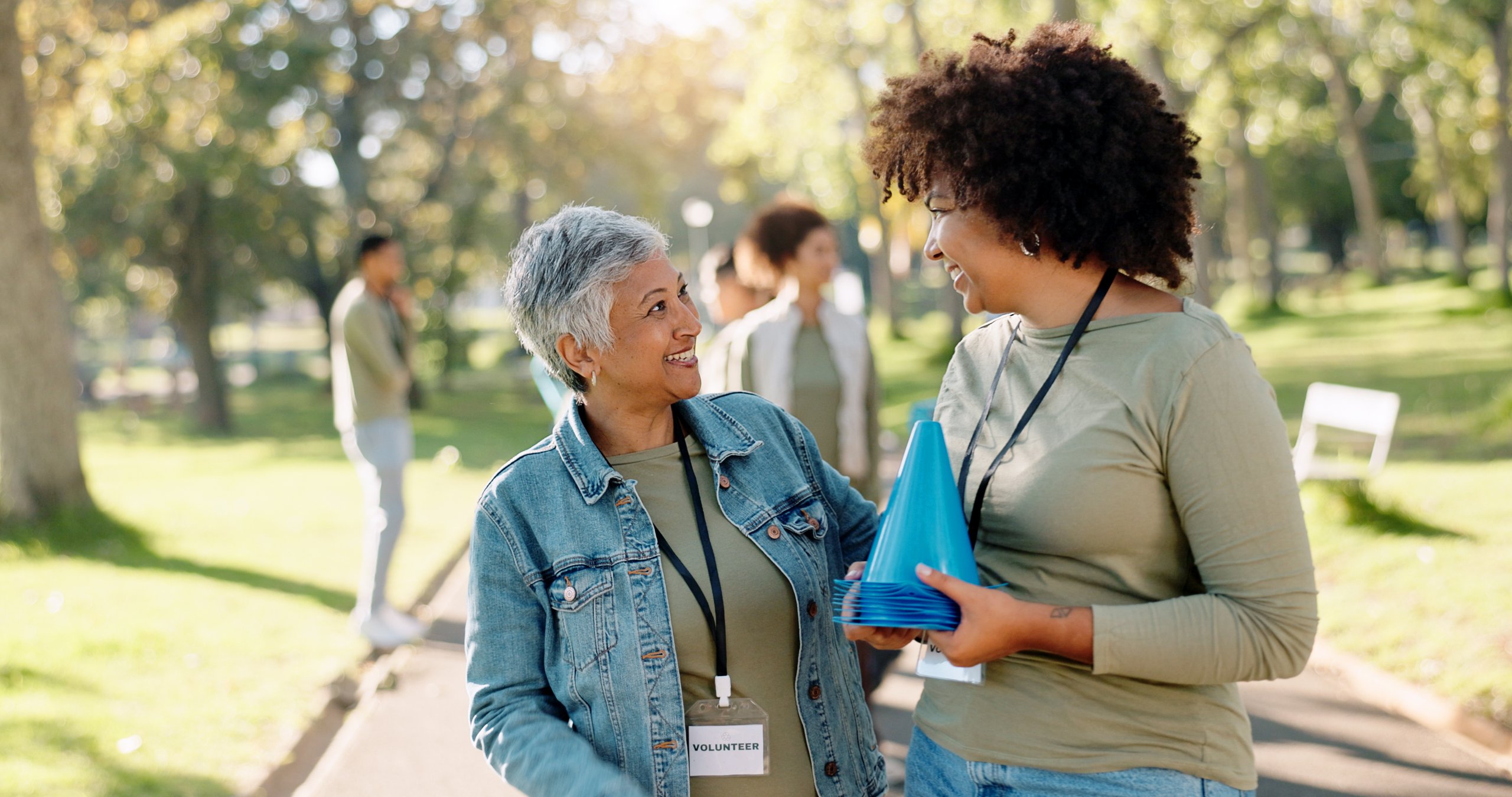 Two women wearing volunteer badges smiling and holding blue cones in a sunny park setting.