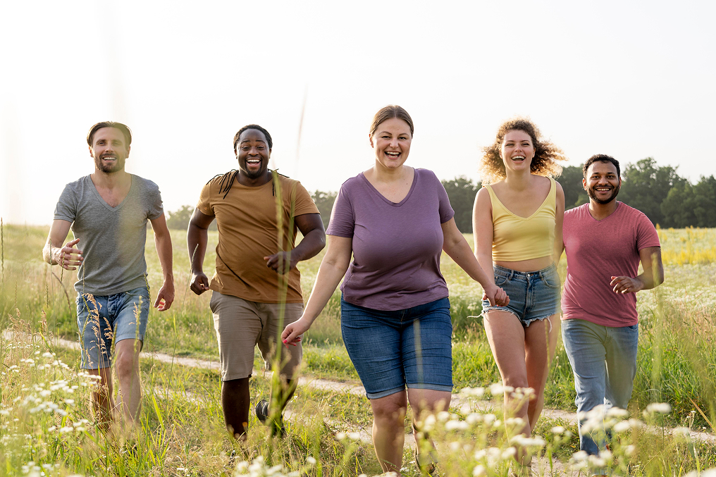 Group of five diverse young adults smiling and running toward the camera on a grassy path in a sunny field.
