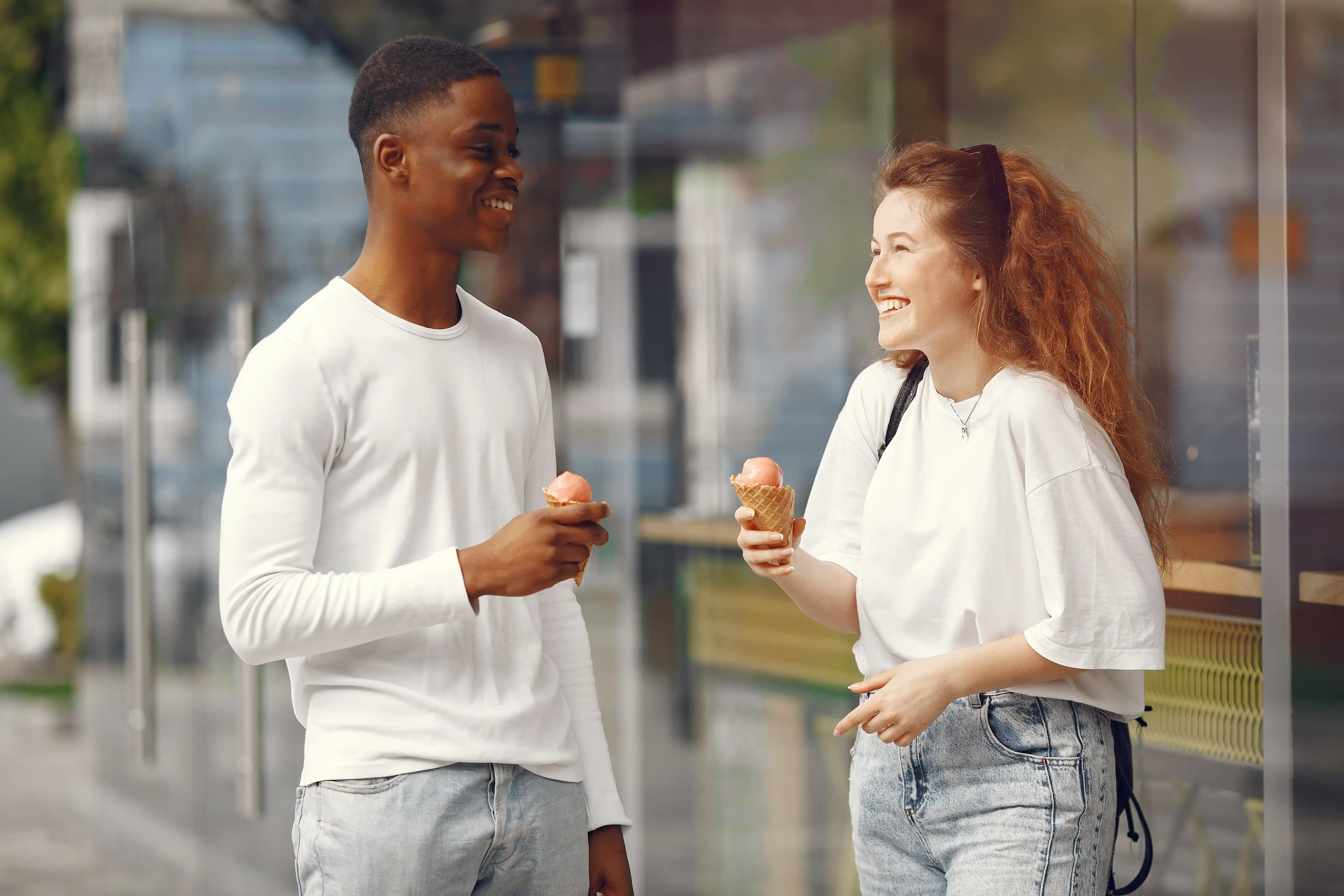 Smiling young interracial couple wearing white tops enjoying ice cream cones outside.