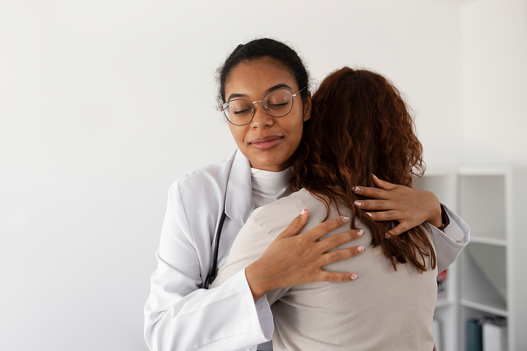 Female doctor wearing glasses and white coat comforting and hugging a patient in a bright office.
