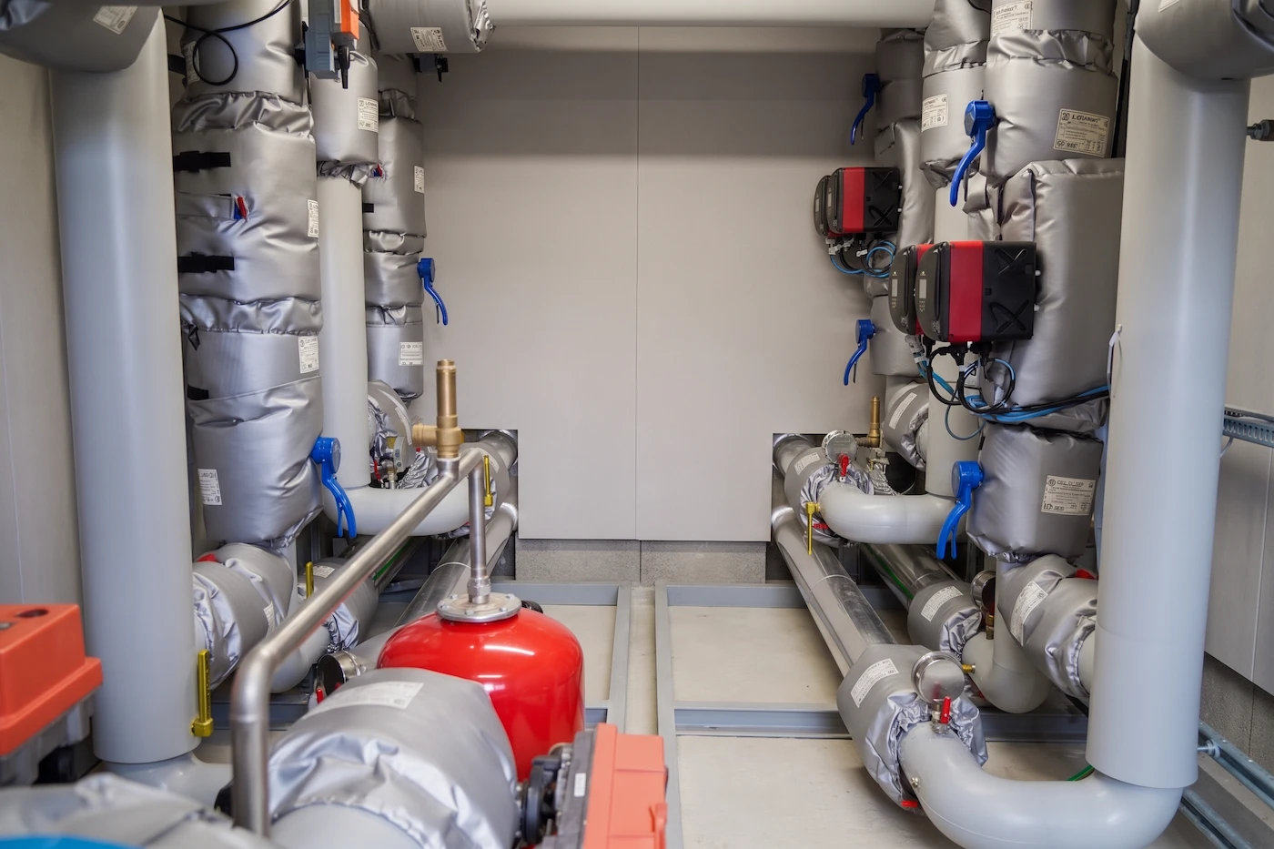 Salle technique avec tuyaux isolés gris disposés le long des murs, vannes bleu vif, appareils de contrôle électroniques rouges et un réservoir rouge au sol.
