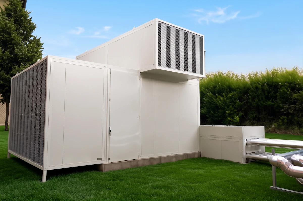 Une unité industrielle blanche pour la climatisation ou la ventilation installée sur une pelouse verte, avec un ciel bleu clair et des arbustes en arrière-plan.