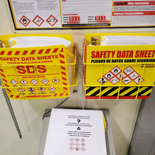 An OSHA-compliant SDS safety station provided by A/F Business Solutions, featuring bright yellow wall-mounted Safety Data Sheet binders, hazard communication posters with GHS pictograms, and workplace emergency contact details.