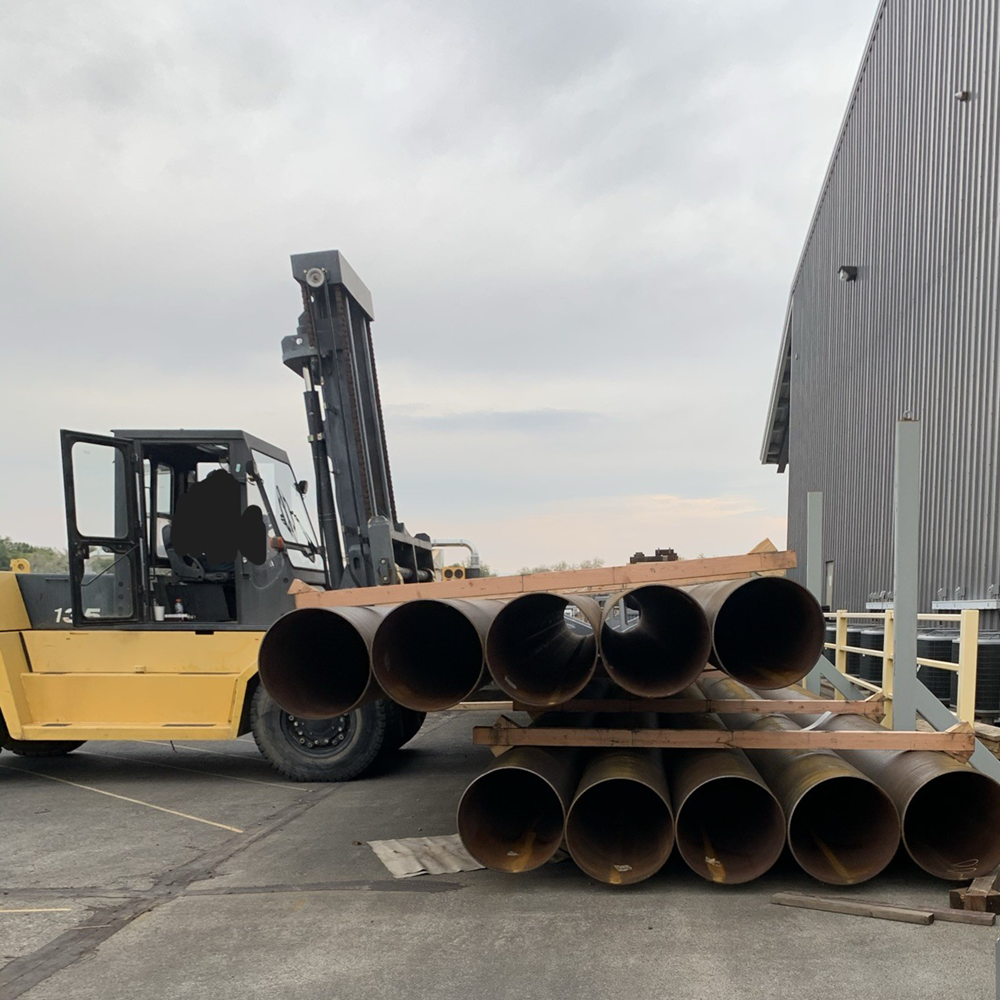 A large Hyster forklift carrying stacked steel pipe outside a manufacturing facility.