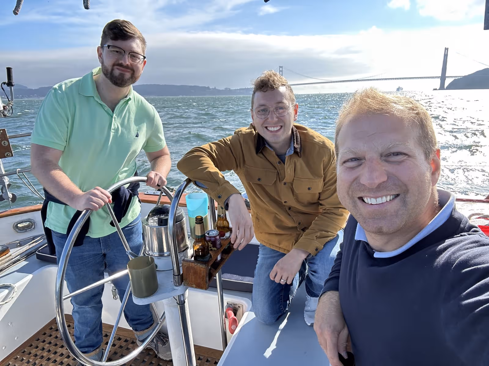 Three men smiling on a boat with the Golden Gate Bridge and water in the background on a sunny day.