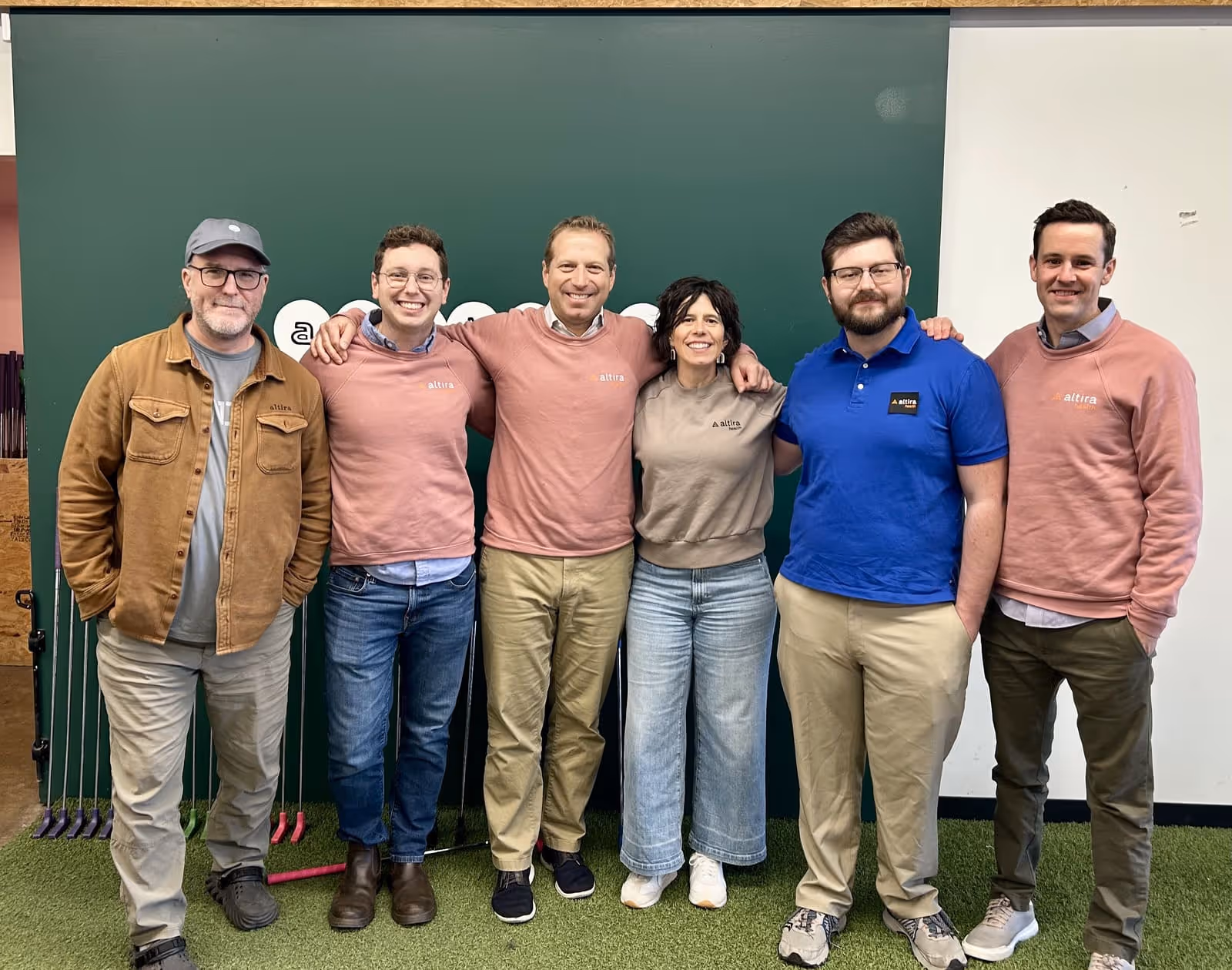 Six people standing closely together indoors on artificial grass, smiling at the camera in front of a green and white wall.