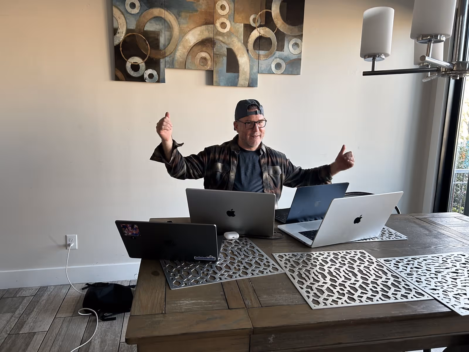 Man wearing glasses and a backward cap sitting at a table with four open laptops giving a thumbs-up with both hands.