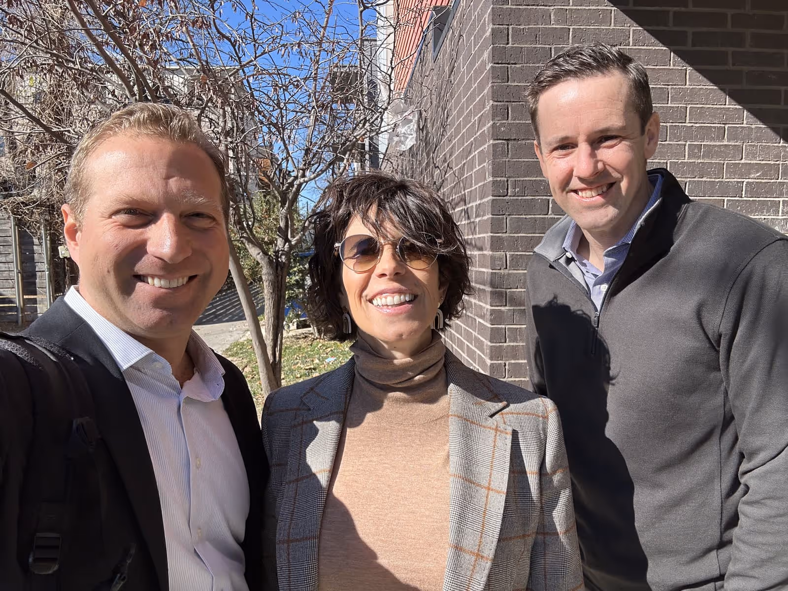 Three people smiling outdoors in sunlight, standing by a brick wall and leafless tree.