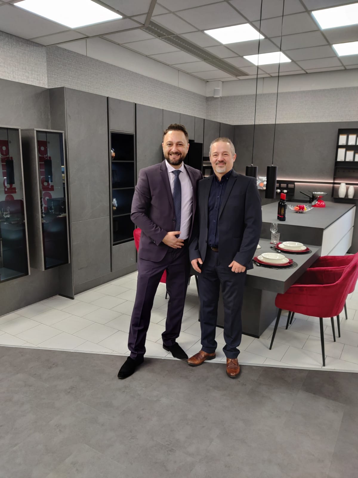 Zwei Männer in Anzügen stehen lächelnd in einer modernen Küche mit roten Stühlen und gedecktem Tisch.