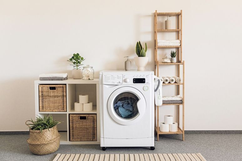 Weiße Waschmaschine in minimalistischem Waschraum mit Regalen, Pflanzen, Handtüchern und Toilettenpapier.