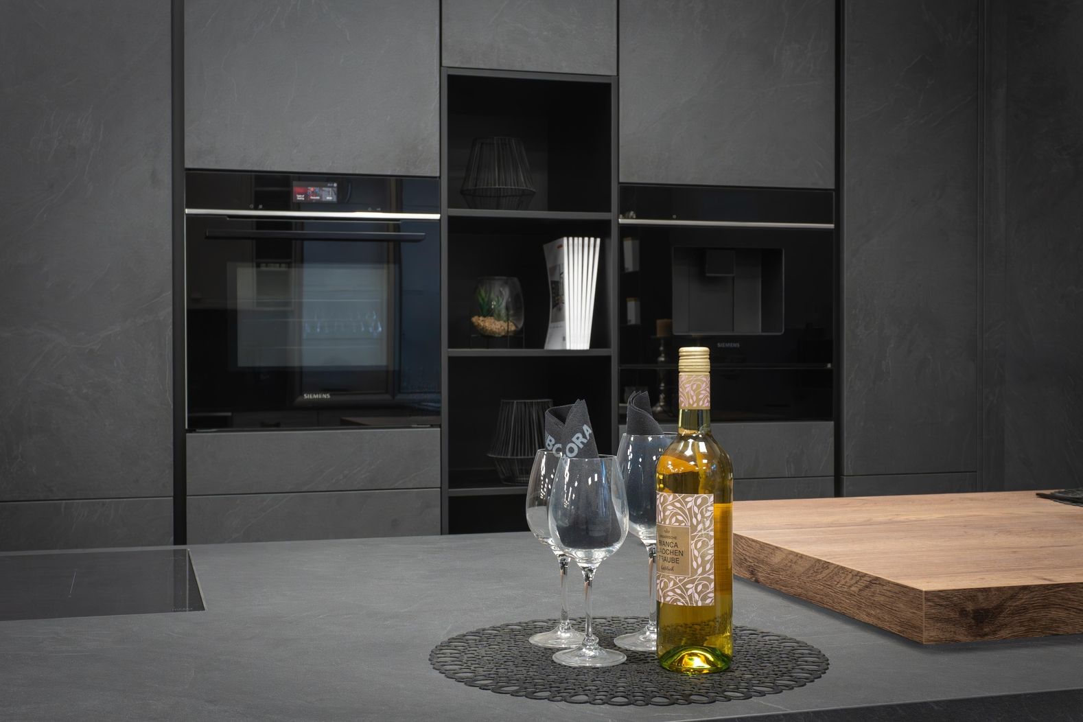 Moderne Küche mit grauen Schränken, einem Flaschen Weißwein und drei Weingläsern mit schwarzen Servietten auf der Arbeitsplatte.