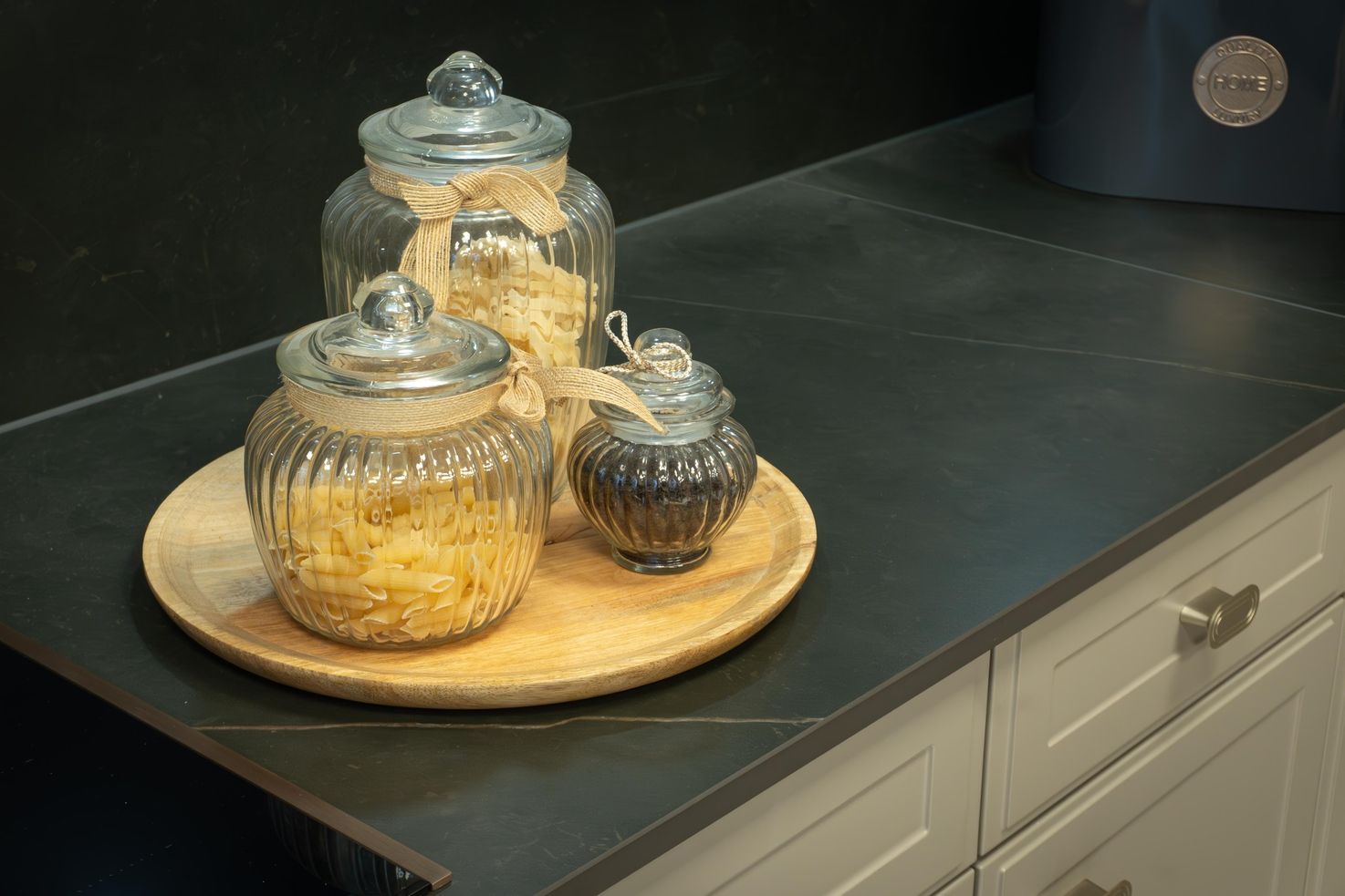 Drei Glasbehälter mit Deckeln, gefüllt mit Pasta und Gewürzen, auf einem Holztablett auf einer schwarzen Küchenarbeitsplatte.