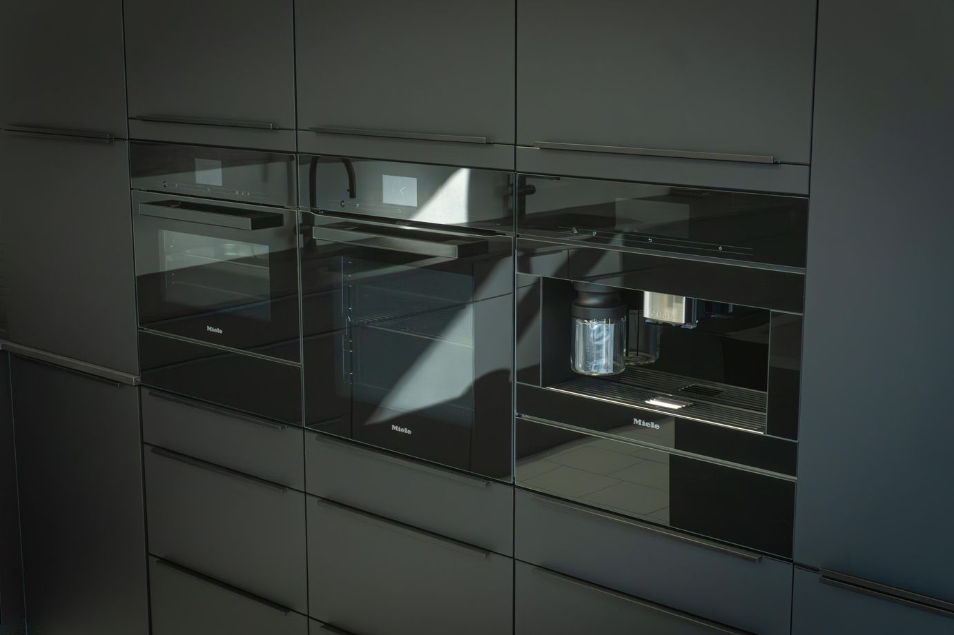 Moderne schwarze Einbauküche mit drei Miele Geräten, darunter zwei Backöfen und eine Kaffeemaschine.
