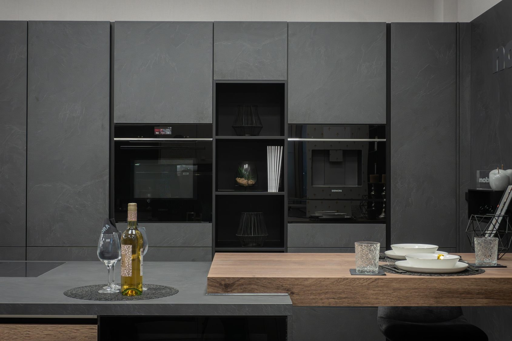 Moderne Küche mit grauen Schränken, eingebauten Siemens-Geräten, einer Flasche Weißwein und Gläsern auf der Arbeitsplatte sowie gedecktem Holztisch.