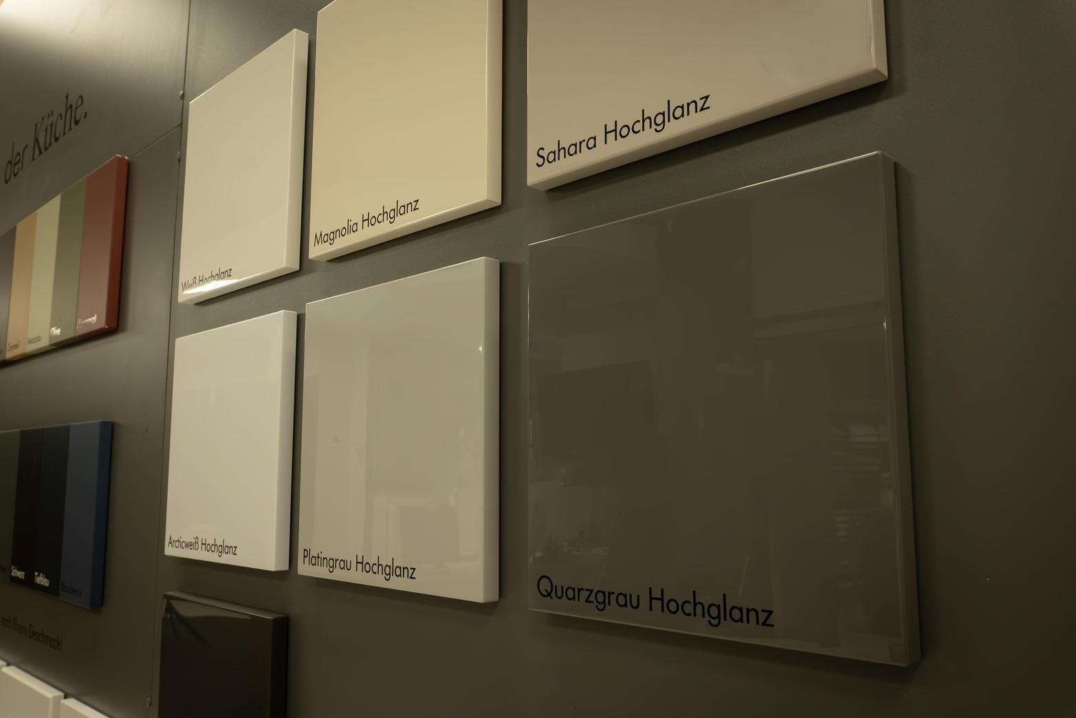 Sechs glänzende Farbmusterplatten an einer Wand, darunter Weiß Hochglanz, Magnolia Hochglanz, Sahara Hochglanz, Arcticweiß Hochglanz, Platingrau Hochglanz und Quarzgrau Hochglanz.