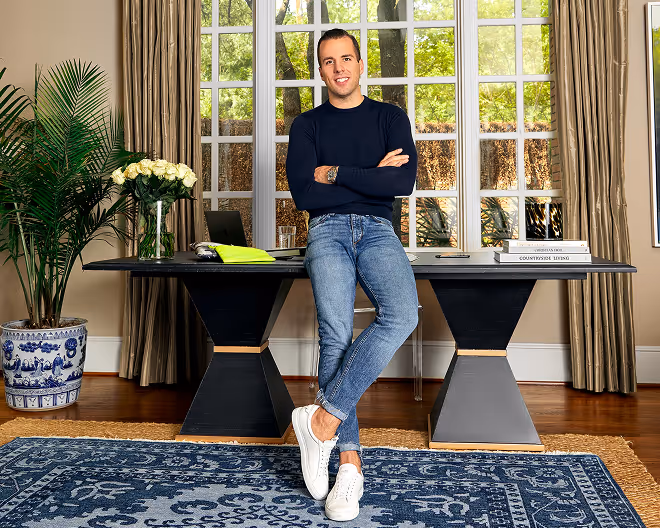 Man in a navy sweater and jeans leaning against a modern desk in a room with large windows, plants, and a blue patterned rug.