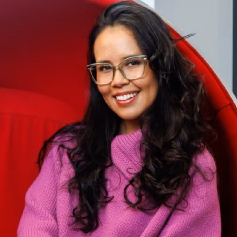 Smiling woman with long curly hair wearing glasses and a purple sweater sitting in a red chair.