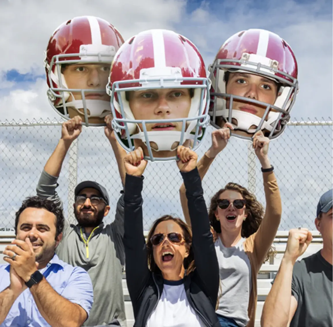 Group of fans cheering at a sports event with three holding large cutouts of football players' heads wearing red helmets.