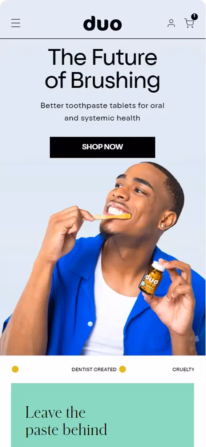 Smiling man in blue shirt brushing his teeth with a wooden toothbrush while holding a bottle of duo toothpaste tablets.