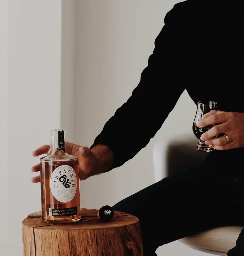 Person holding a glass of Oakpacker whiskey beside the bottle on a wooden stump