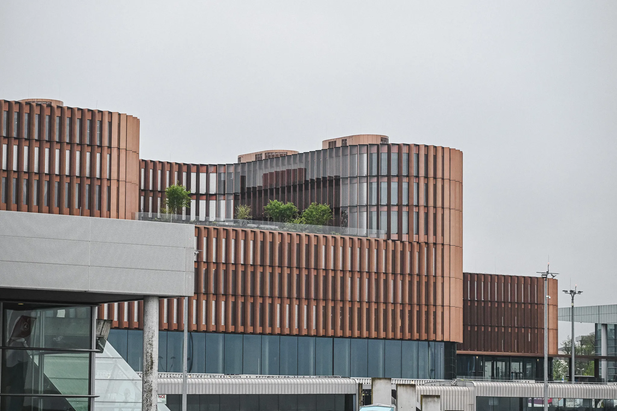 Modern building with curved facade and vertical slats, featuring rooftop greenery under a gray sky.
