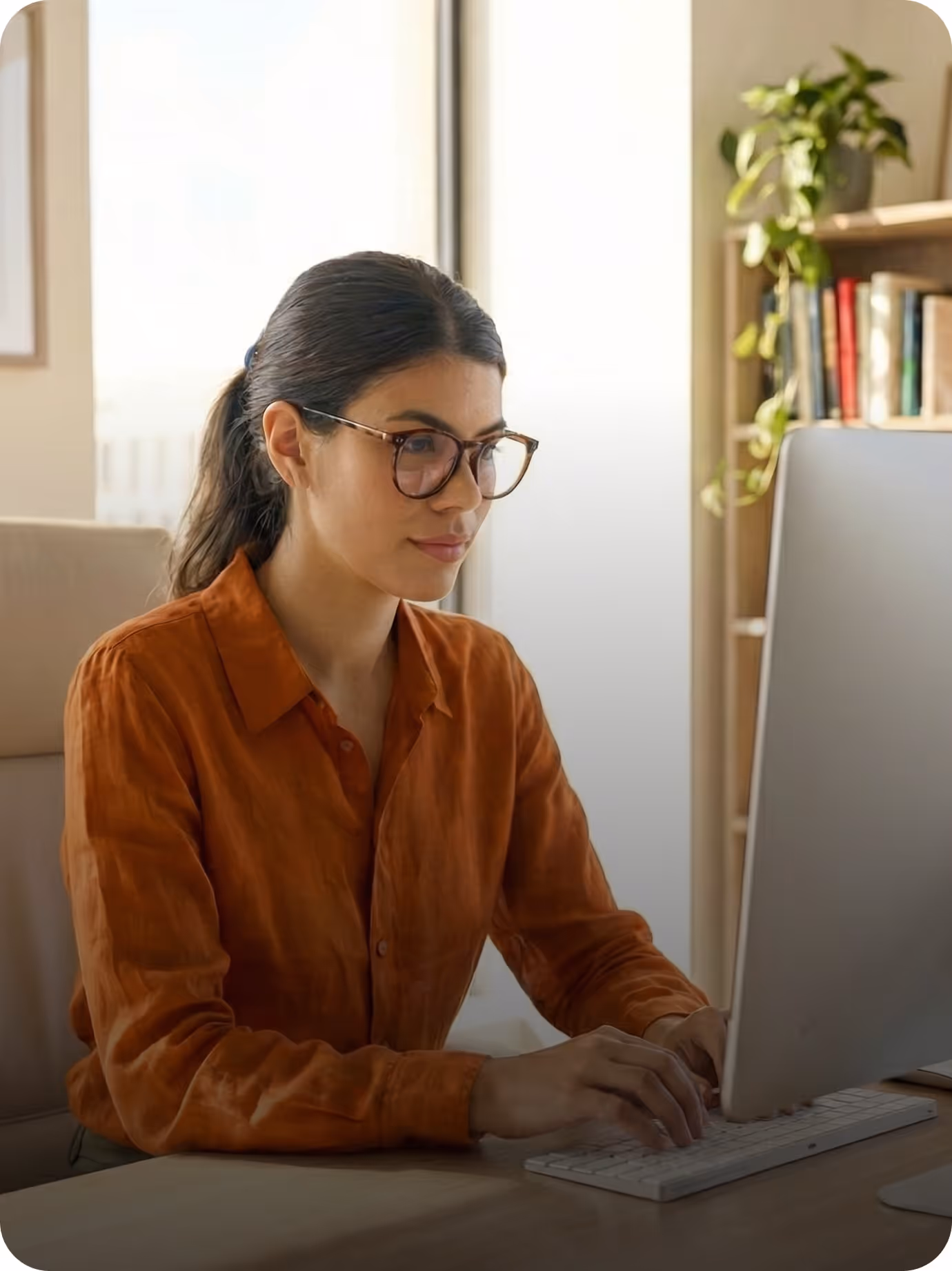 Woman with glasses in orange shirt working on a desktop computer at a desk in a home office.