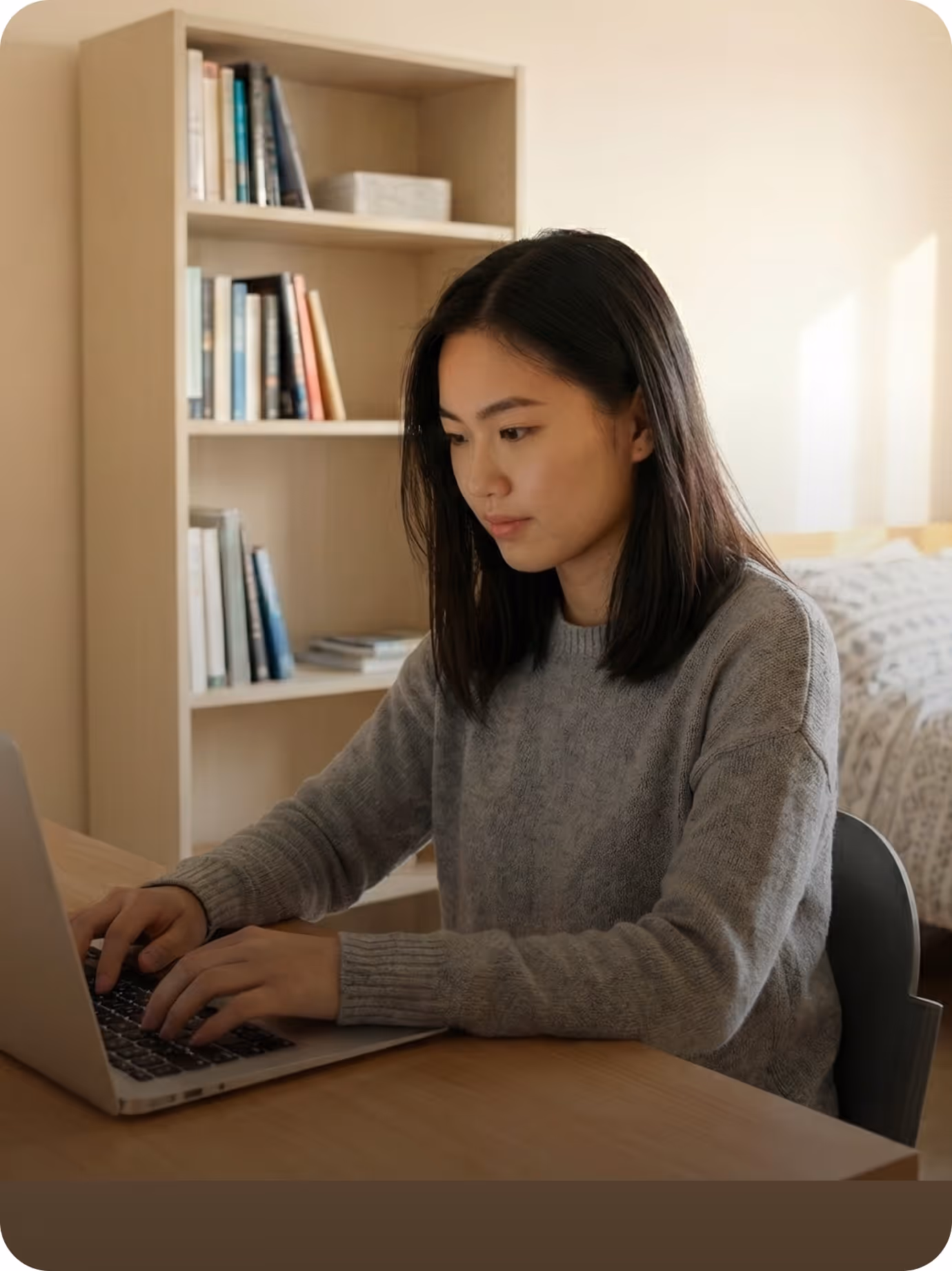 Young woman wearing gray sweater types on a laptop at a wooden desk in a cozy room.