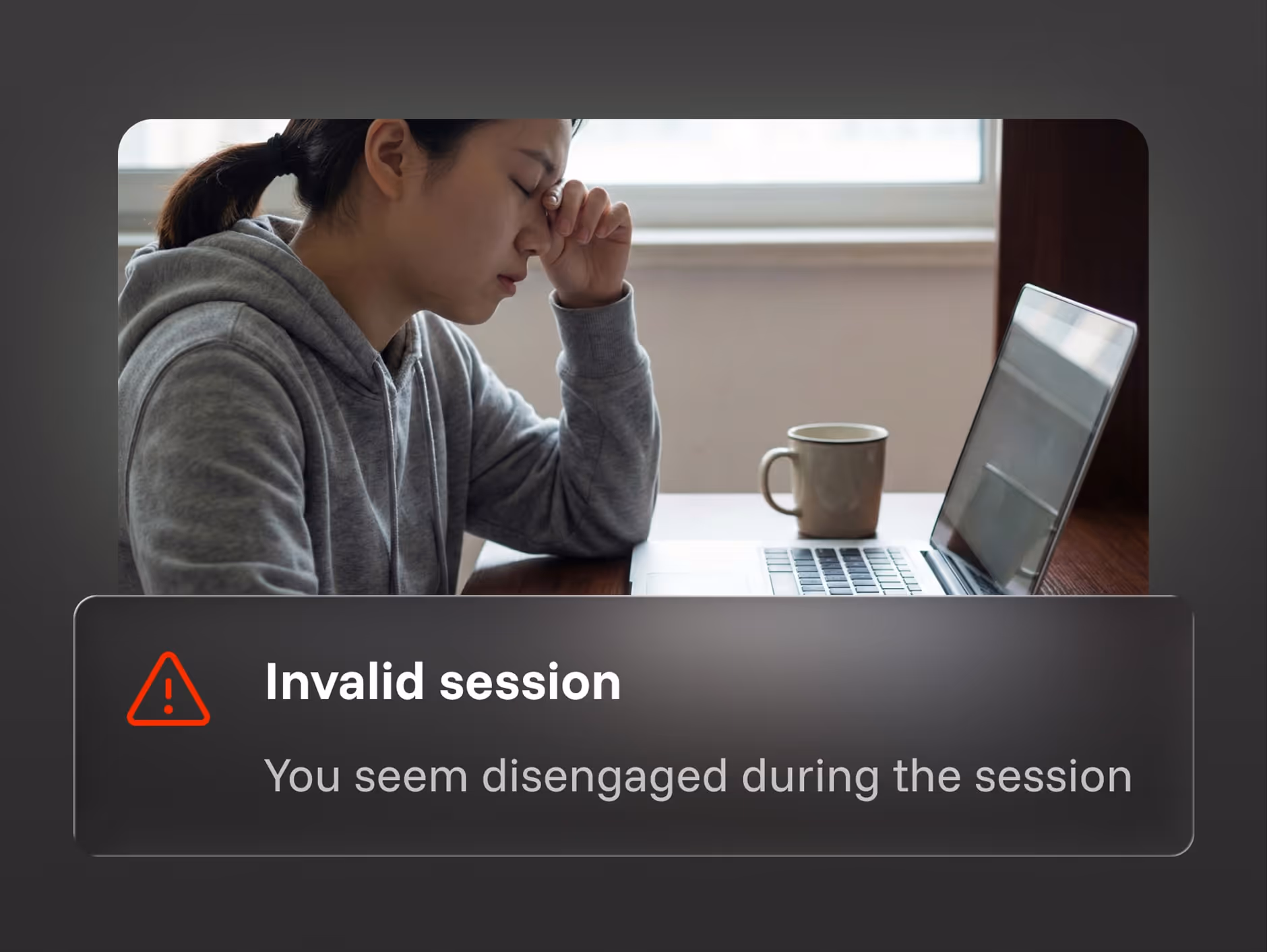 Tired woman in gray hoodie sitting at a desk with laptop and mug, eyes closed, pinching nose bridge.