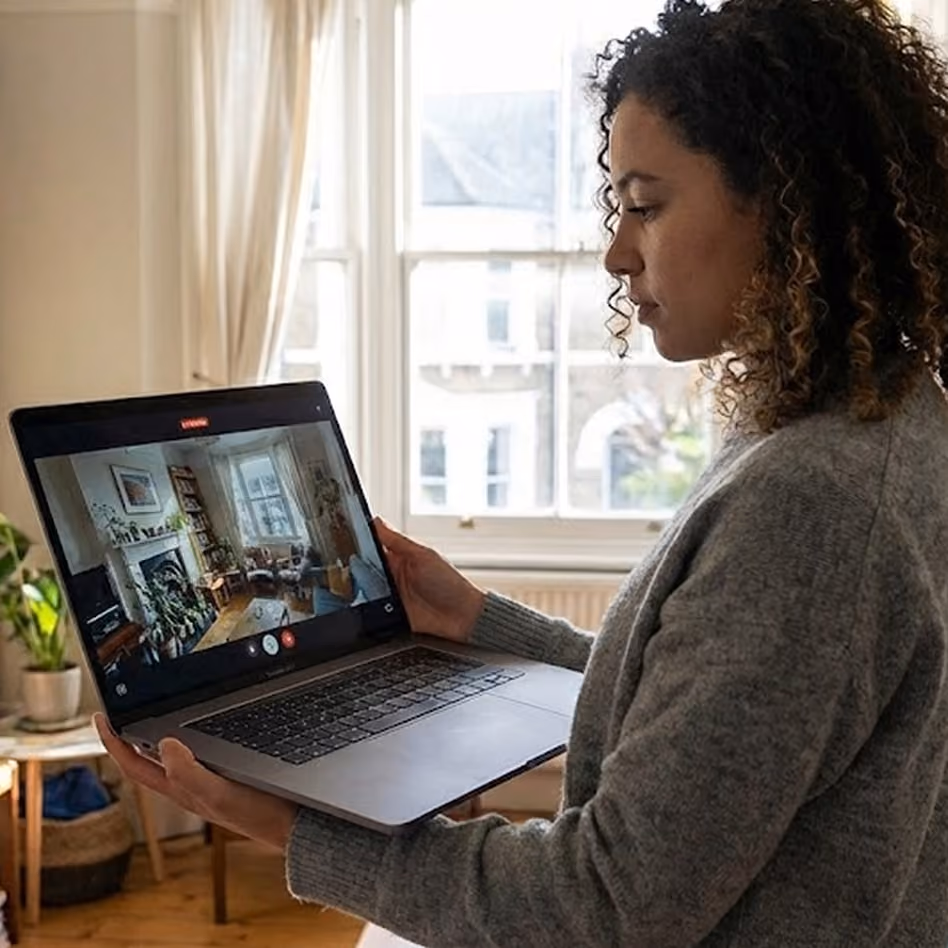 Woman holding a laptop showing a room scan with furniture and plants.