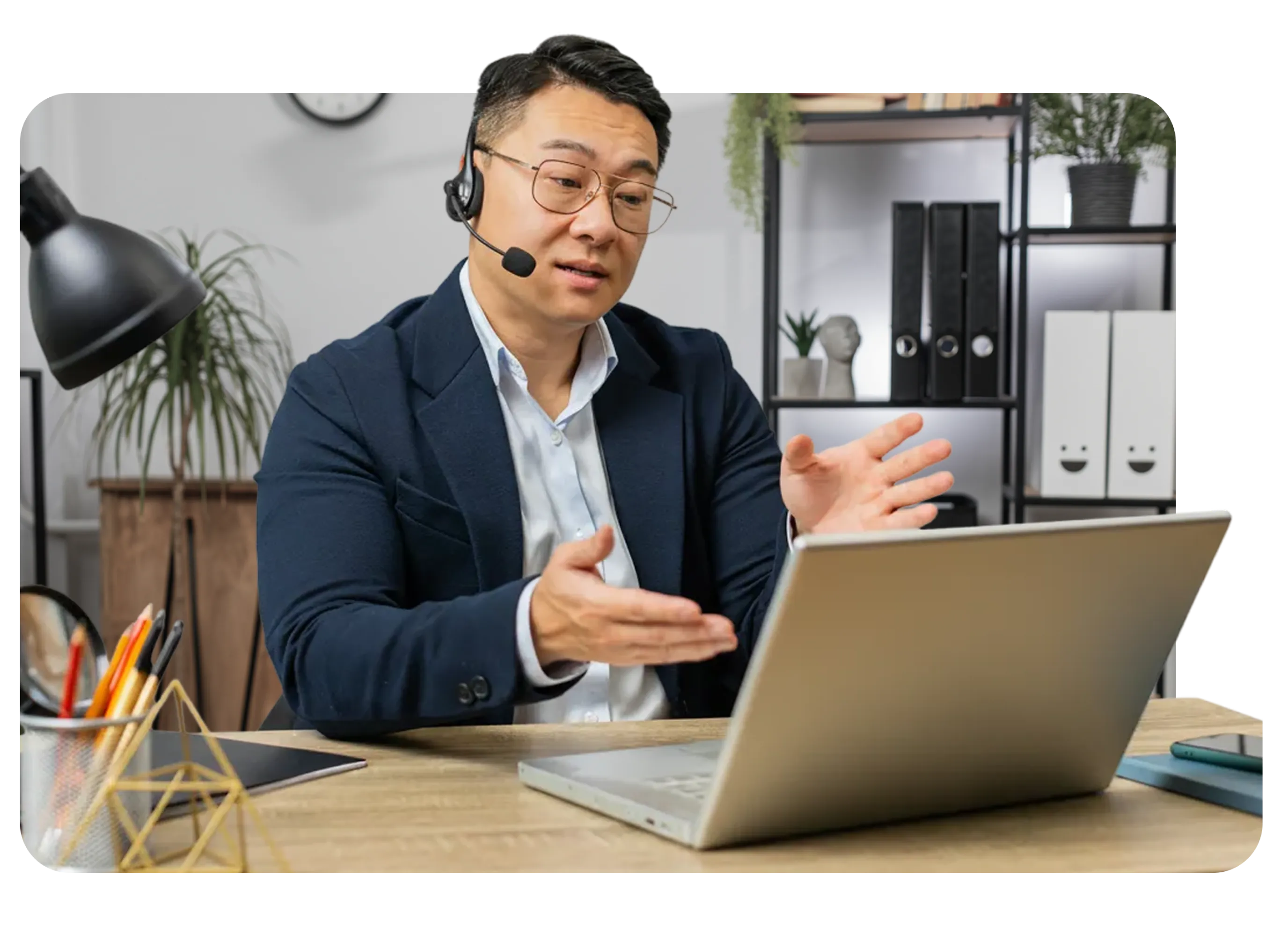 Man in glasses and headset gesturing while speaking during a video call at a desk with a laptop.