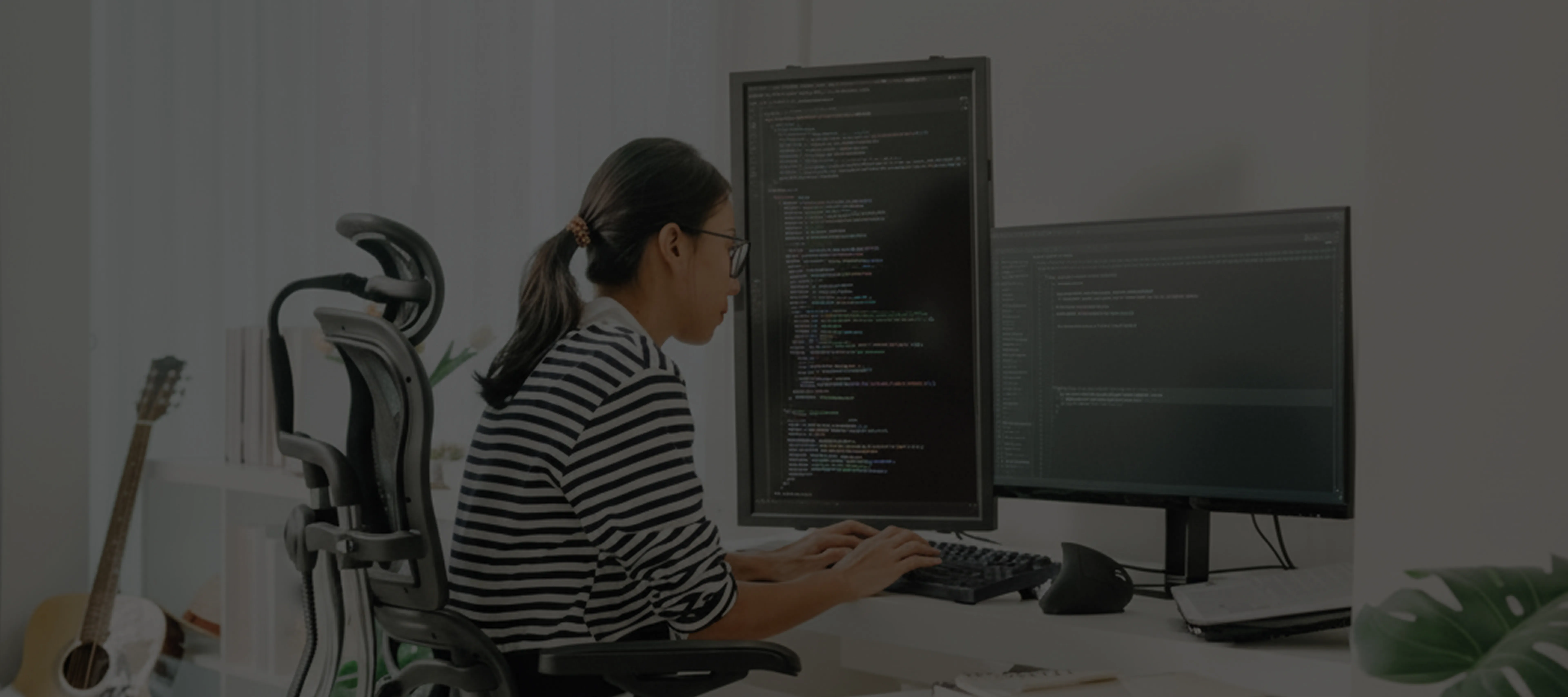 Person in glasses and striped shirt typing on keyboard with two monitors displaying code.