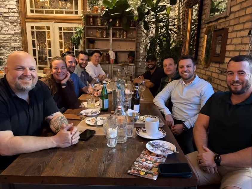 Eight people smiling and sitting around a wooden table in a restaurant with drinks and plates.