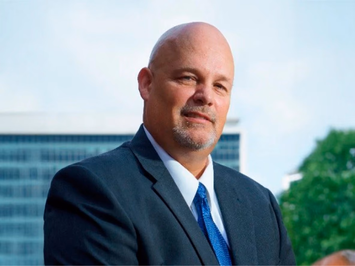 Man in a suit with blue tie standing outdoors with a building and trees in the background.