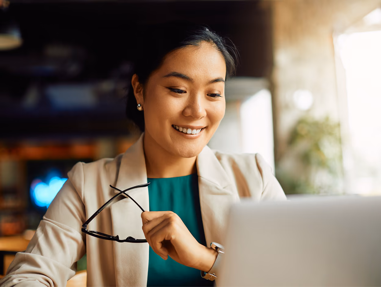 Smiling woman holding glasses looking at a laptop in a softly lit indoor setting.